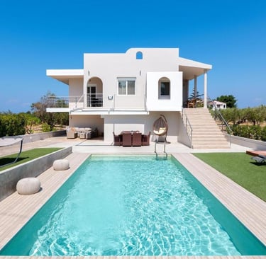 Luxury white villa with a turquoise swimming pool, outdoor dining, and lounge area under a clear blue sky.