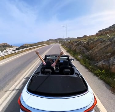 A white Volkswagen convertible driving along a scenic coastal mountain road with the driver's hands raised.