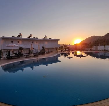Luxury resort swimming pool reflecting a golden sunset over Mediterranean hills and white villas.