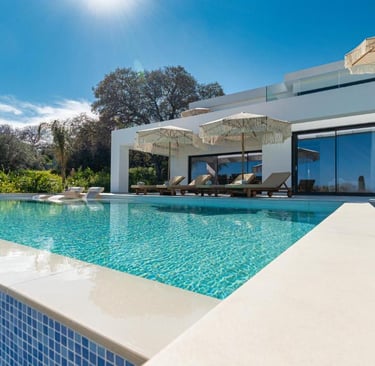 Luxury modern villa with a blue infinity pool, lounge chairs, and sun umbrellas under a clear sky.