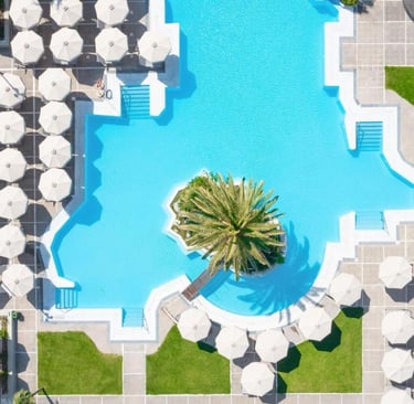Aerial view of a luxury resort swimming pool with white umbrellas and a central palm tree island.