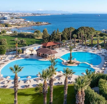 Aerial view of a luxury Greek island resort with a blue swimming pool and palm trees overlooking the Mediterranean Sea.