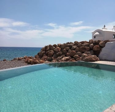 Luxury infinity pool overlooking the Mediterranean Sea with a white Greek chapel and rocky coastline.
