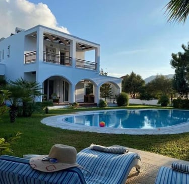 Luxury white villa with private swimming pool, garden, and sun loungers under a blue sky.