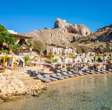 Luxury beach club in Lindos, Rhodes, featuring sun loungers and cabanas under the historic Acropolis cliff.