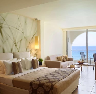 Luxury beachfront hotel room with king bed, lounge seating, and private balcony overlooking the ocean.