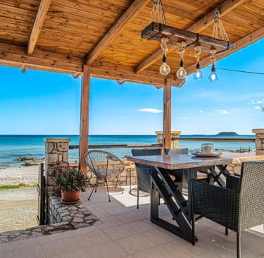 Rustic outdoor patio with dining table and wood pergola overlooking a Mediterranean beach.