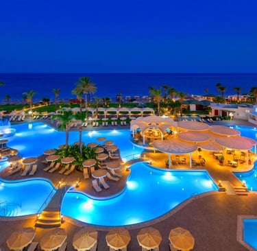 Luxury beach resort at night featuring illuminated swimming pools and palm trees by the ocean.