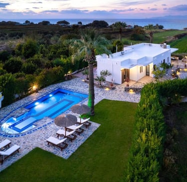 Aerial view of a luxury white villa with a private pool and sun loungers at sunset near the ocean.