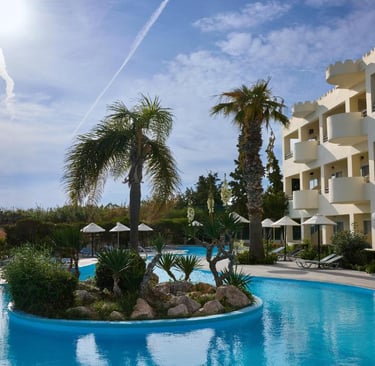 Luxury tropical resort swimming pool with palm trees and hotel balcony views under a sunny sky.