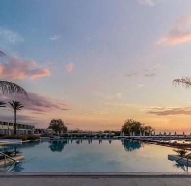 Luxury resort swimming pool reflecting a vibrant sunset sky with palm trees and lounge chairs.