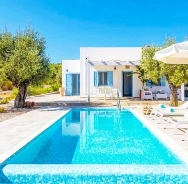 Luxury white Greek villa with a private swimming pool, sun loungers, and olive trees under a clear blue sky.