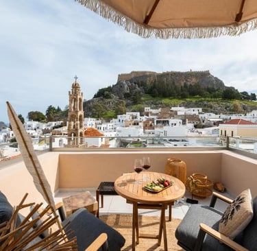 Luxury balcony view of Lindos Acropolis in Rhodes, Greece, with wine and snacks on a patio table.