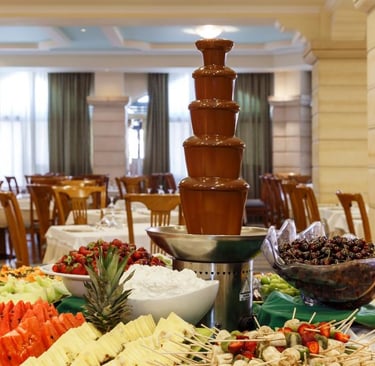 A tiered chocolate fountain on a buffet table surrounded by fresh fruit platters in a hotel restaurant.
