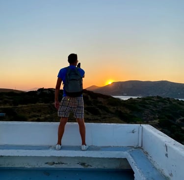 A male traveler with a backpack takes a sunset photo from a rooftop overlooking mountains and the sea.
