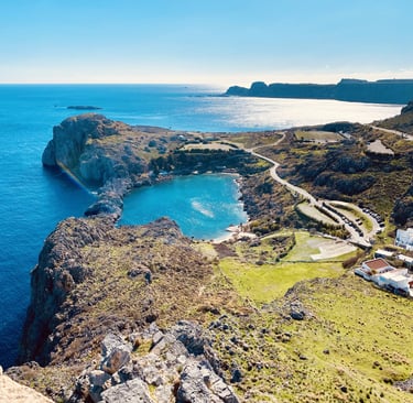 Aerial view of St. Paul's Bay in Lindos, Rhodes, featuring turquoise water, rocky cliffs, and the Mediterranean Sea.