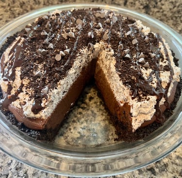 A homemade chocolate silk pie with a sliced section, topped with whipped cream, cookie crumbles, and syrup.