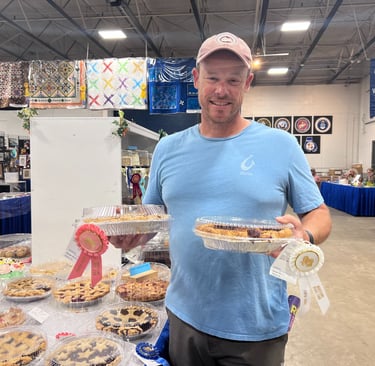 A man smiling at a county fair home arts competition holding award-winning baked fruit pies with ribbons.