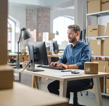 Hombre sentado trabajando en una computadora en una oficina. Fondo de estanterias y cajas apiladas.