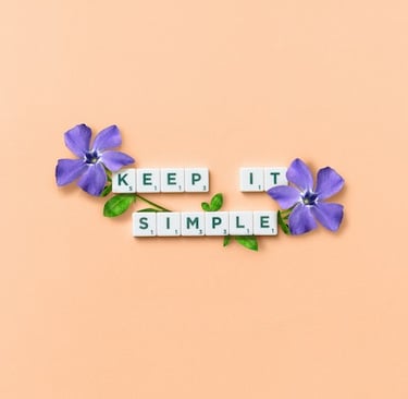 Scrabble tiles spelling out the words keep it simple on an orange background with 2 purple flowers