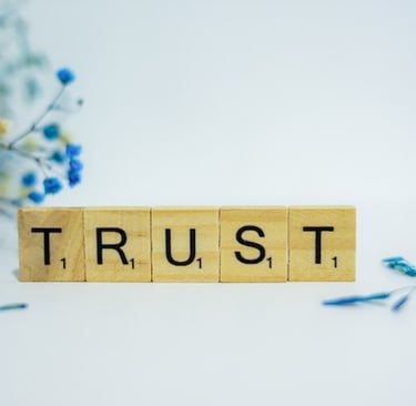 Scrabble tiles spelling out the word trust with some out-of-focus blue flowers in the background
