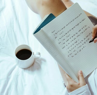 Imagem de um caderno com algumas coisas escritas, uma mão segurando uma caneta e uma caneca de café 