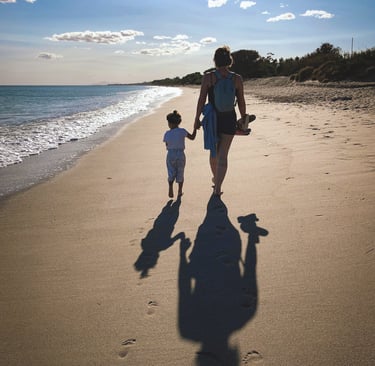 mother holds toddler's hand as they walk along a beach