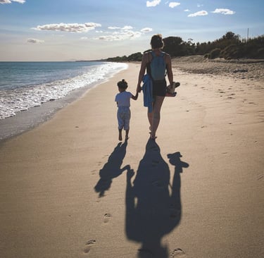 mother holds toddler's hand as they walk along a beach