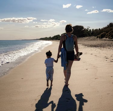 mother holds toddler's hand as they walk along a beach