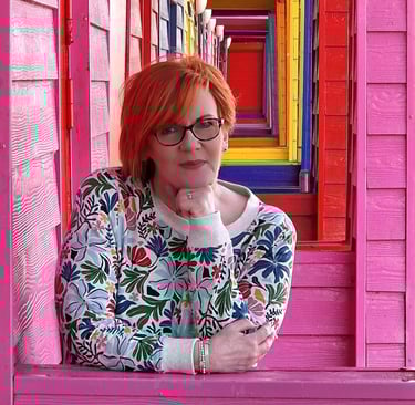 Dawn Vincent Celebrant leaning on a pink beach hut with a row of colourful huts in the background