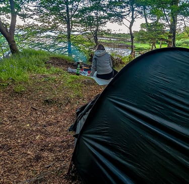 Campement en forêt en Écosse, avec une tente noire et une personne assise près d'un feu, au bord d'un lac.