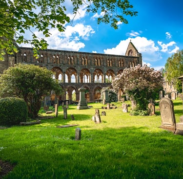 Ruines majestueuses de l'Abbaye de Jedburgh sous un ciel bleu, entourées d'arches gothiques, verdure et pierres tombales.