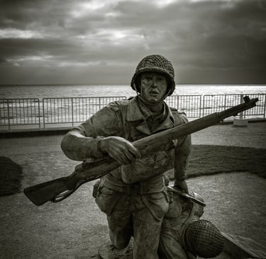 Statue en bronze d'un soldat courant avec un fusil, traînant un camarade tombé. Mer, obstacles et ciel nuageux en fond.