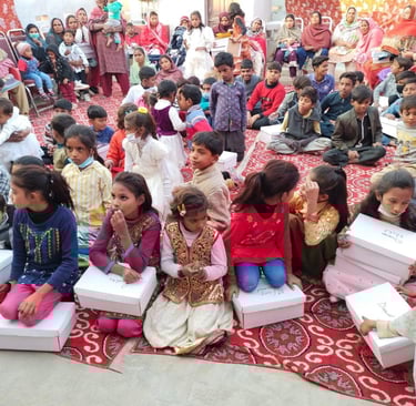A group of children sitting on a red patterned rug outdoors, holding gift boxes during a community event.