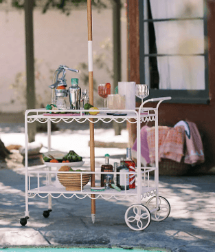 A white vintage-style bar cart with summer cocktails and snacks by a swimming pool under a green striped umbrella.