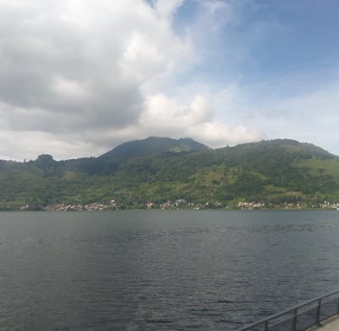 a mountain in the background is getting covered in clouds with houses at the bottom of the mountain by on a lake
