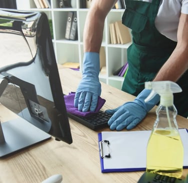 Man wiping down keyboard while wearing cleaning gloves, green apron. Office setting in the background with cleaning supplies