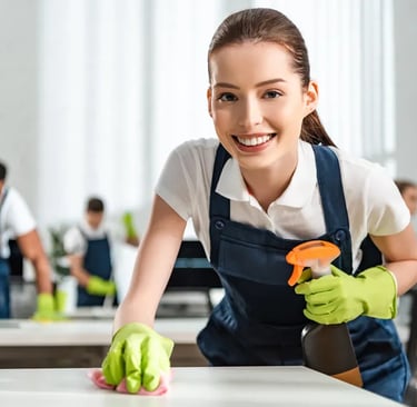 Smiling brunette woman wearing navy apron, green cleaning gloves & wiping down desk.  Workers clean in background.
