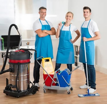 Three smiling people wearing blue aprons ready to clean with mops, cleaning bucket, supplies and an industrial vacuum