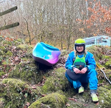 a woman in a dry suit with a white water kayak on a mossy hill 