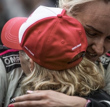 Two female race car drivers in OMP suits embrace to celebrate a victory on the track.