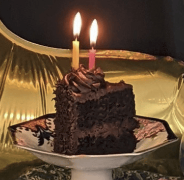 Slice of chocolate cake with burning candles on porcelain trinket tray.