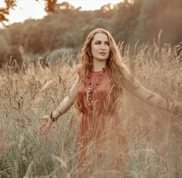 Trauerrednerin Leonie Stratmann steht im altrosanen Sommerkleid in einem Feld mit Wildblumen, hinter ihr Wald.
