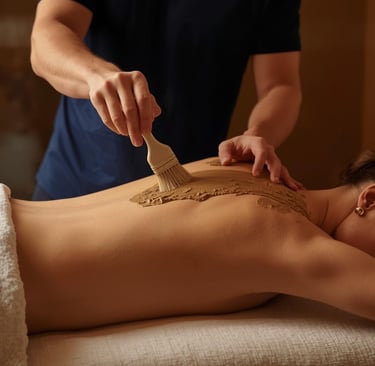 A spa therapist applies a mineral mud mask to a woman's back using a brush during a relaxing body treatment.