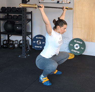 A woman performing an overhead barbell squat in a home garage gym with weight plates.