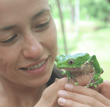 Smiling woman gently holding a large green frog, looking at it with affection. 