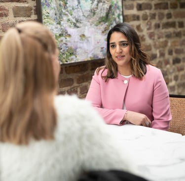 A photo of Anita wearing a pink jacket sitting at a table talking to a person who has their back to the camera.