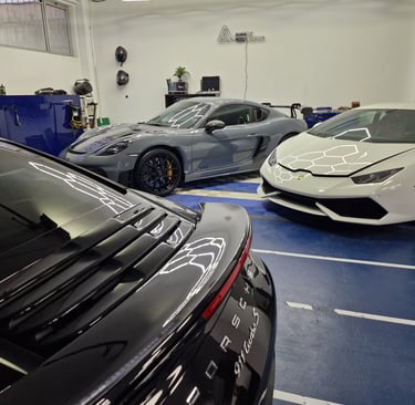 Luxury sports cars including a white Lamborghini and grey Porsche GT4 RS parked inside a modern detailing garage.