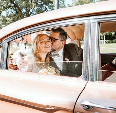 Braut und Bräutigam sitzen glücklich in einem Oldtimer, der Bräutigam küsst die Braut liebevoll auf die Wange, Brautstrauß im