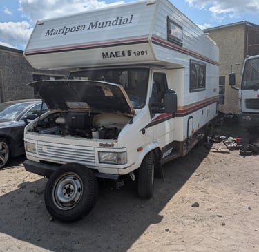  Old garage yard in rural Poland with campervan parked among broken-down cars and engine parts.